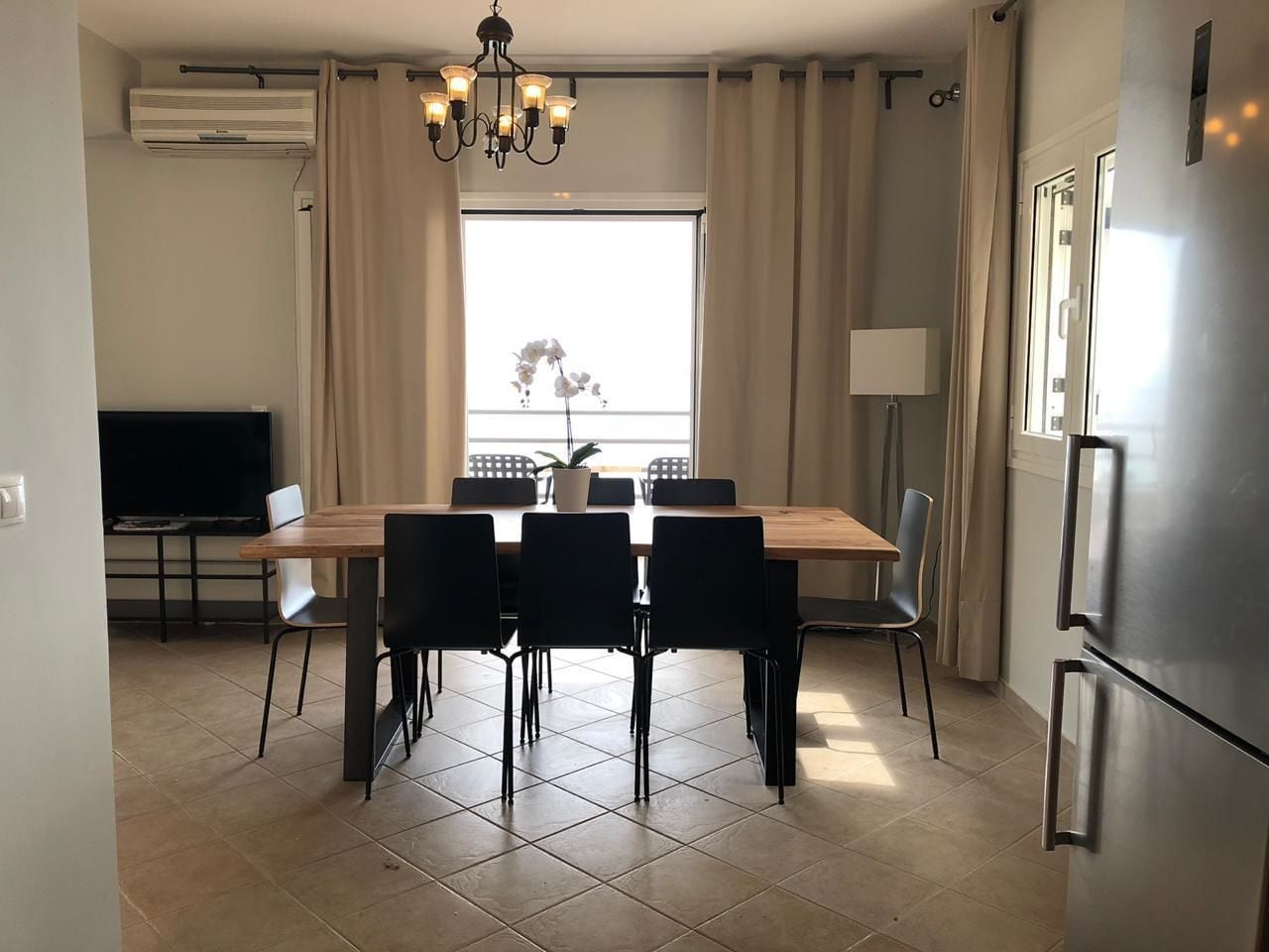 A dining area with a wooden table and chairs, a chandelier, and a large window with curtains.