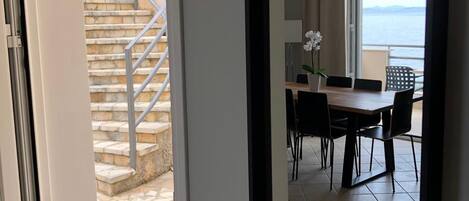A view through a doorway to a dining area with a table, chairs, and a chandelier. A staircase with a metal railing is visible in the background.