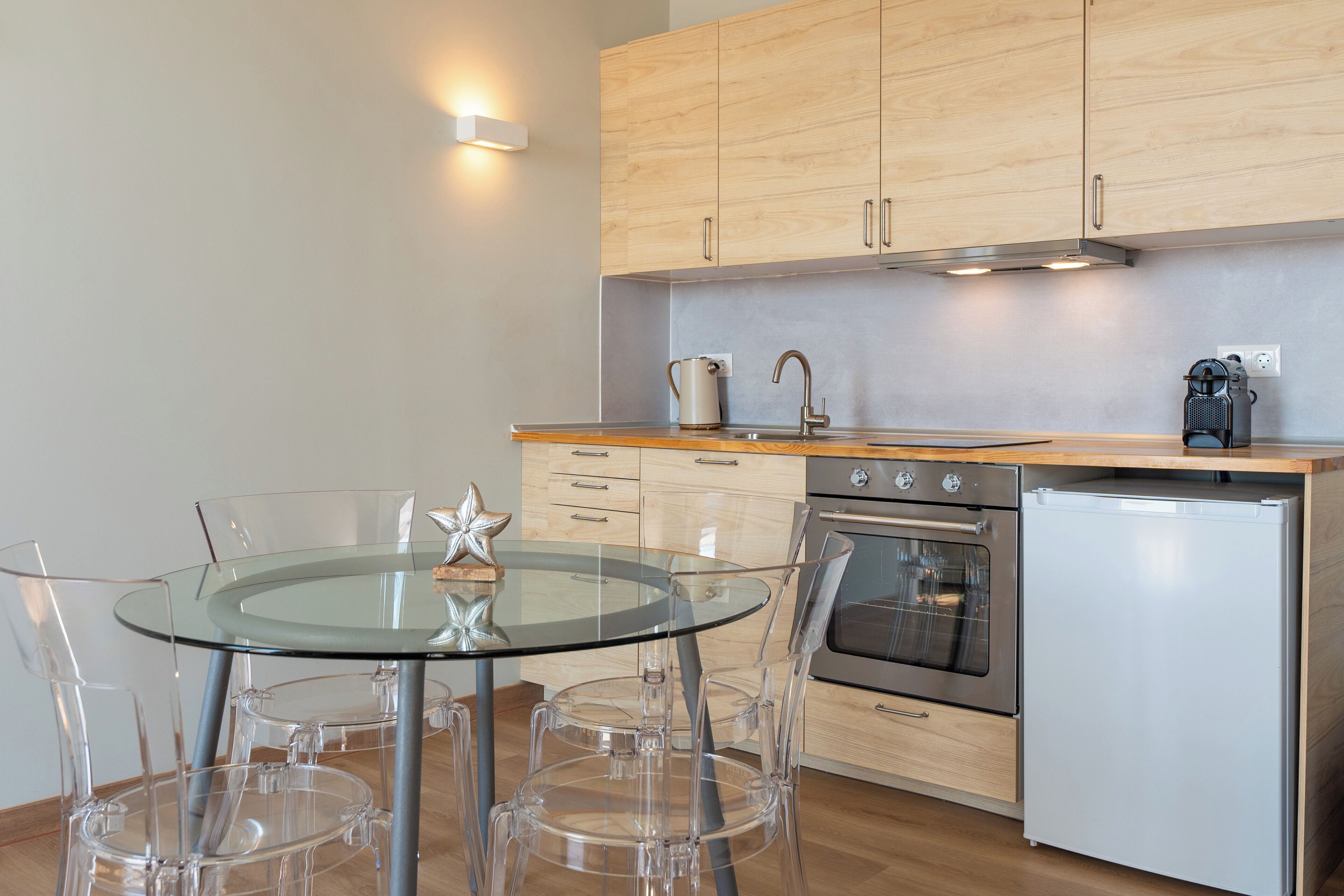 A modern kitchen with wooden cabinets, a stainless steel oven, a white refrigerator, and a glass dining table with clear chairs.
