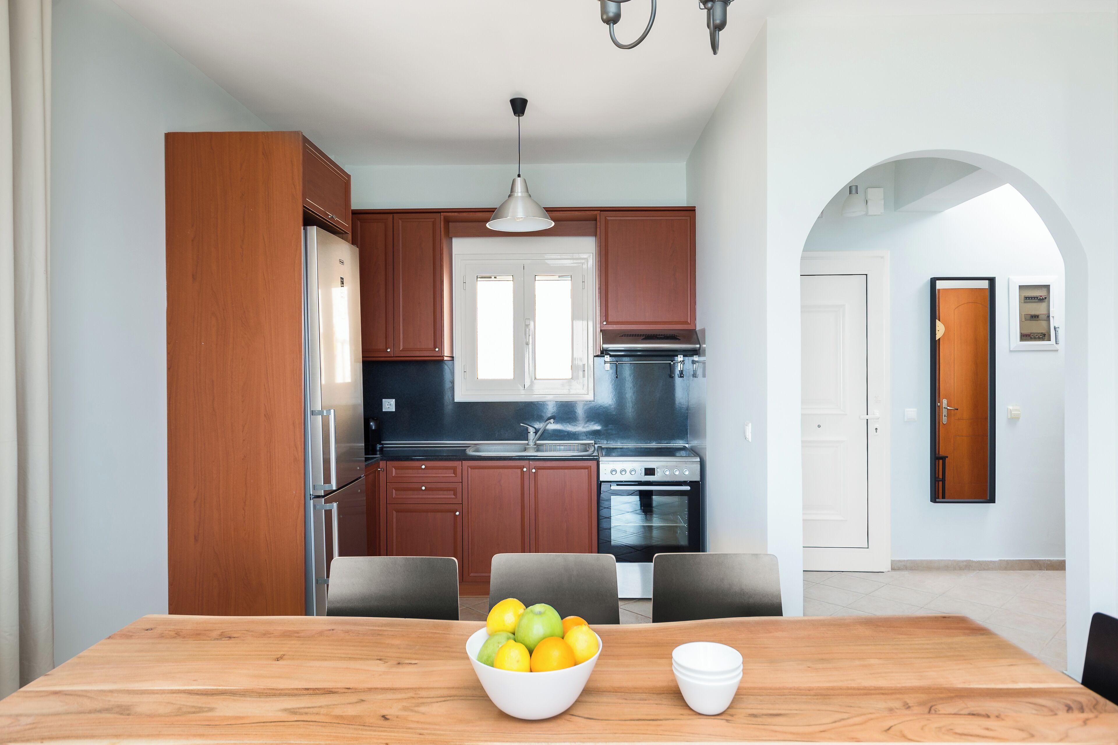 A modern kitchen with wooden cabinets, a stainless steel refrigerator, and a built-in oven. There is a dining table with chairs and a bowl of fruit.