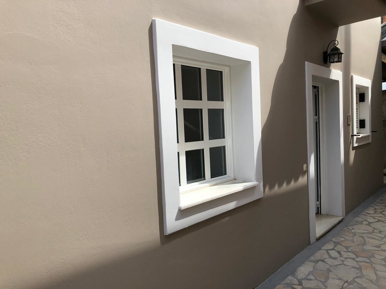 Fachada de edificio con pared color beige, dos ventanas con marco blanco y suelo con diseño de piedra.