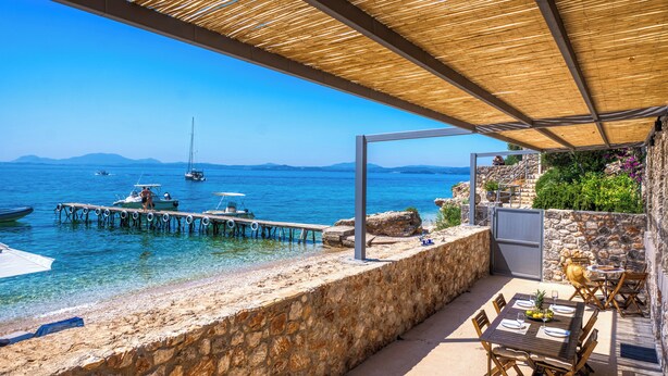 A seaside patio with a stone wall, wooden deck, and outdoor seating area overlooking the sea. - Glyfa Beach Villas (Corfu)