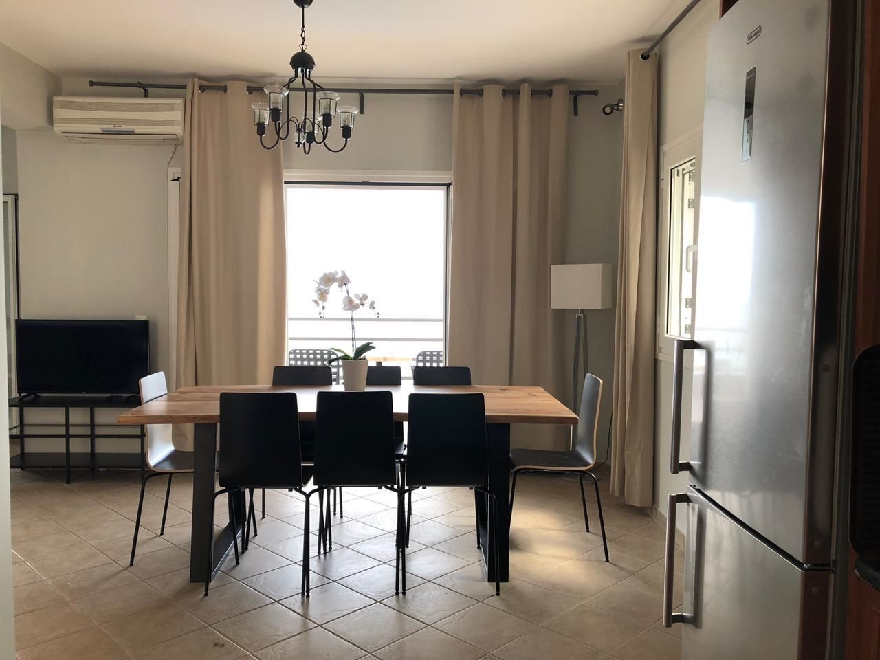 Et moderne køkken med et rustfrit stål køleskab, et spisebord med stole, et fjernsyn og et vindue med gardiner.