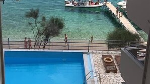 A swimming pool with a stone-tiled deck, a boat docked at the water's edge, and a clear view of the sea. - Glyfa Beach Villas (Corfu)