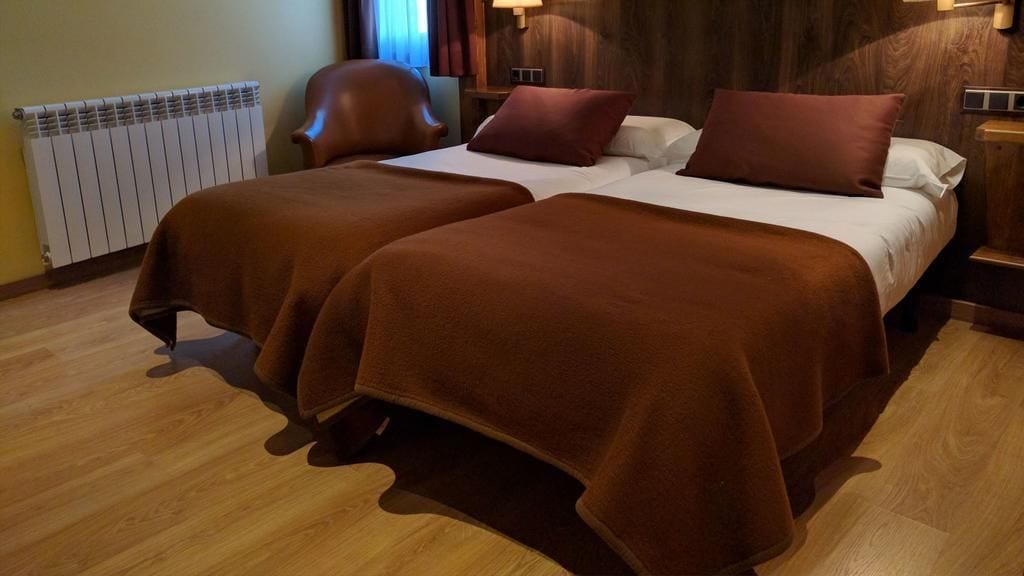 A hotel room with two beds, a wooden floor, a radiator, a chair, and a nightstand with a lamp.
