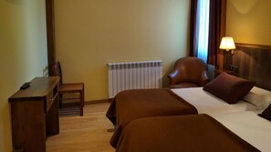 Habitación de hotel con dos camas, un escritorio, una silla, un radiador y una ventana con cortinas.