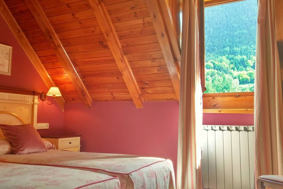 Een slaapkamer met een houten plafond, een bed met bloemenbeddengoed, een nachtkastje, een wandlamp en een raam met uitzicht op een bos.