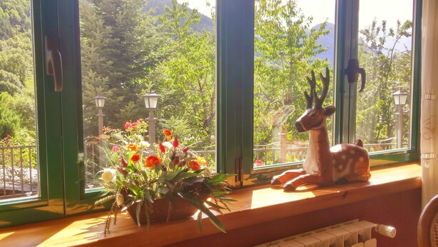 A window with a view of a forested area, a deer statue, and a vase of flowers on a wooden ledge. - Hotel Colomers (Naut Aran)