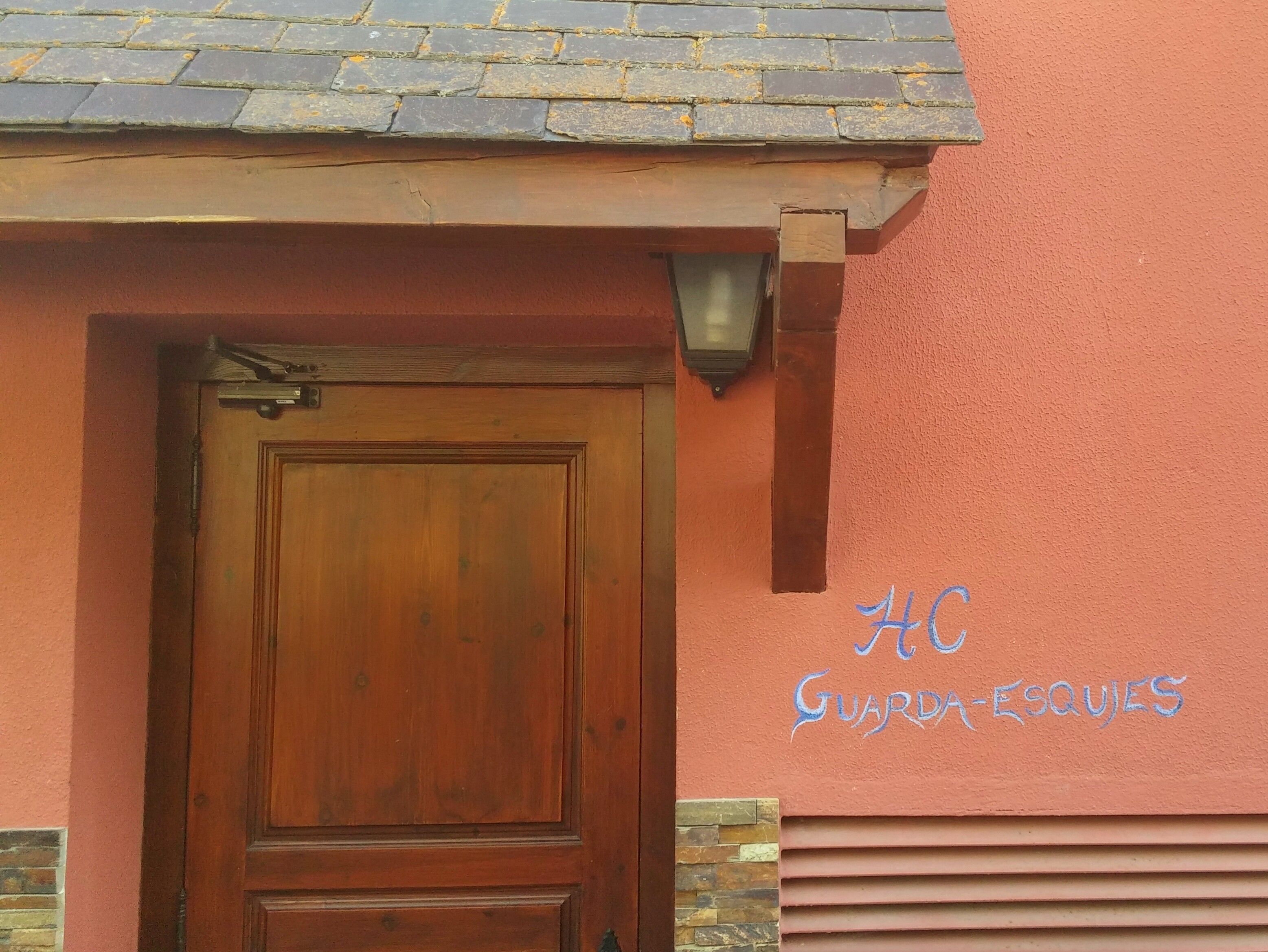 Una puerta de madera con un picaporte metálico, una pequeña ventana encima y una pared con el texto "AC GUARDAS ESQUIES" pintado en azul.