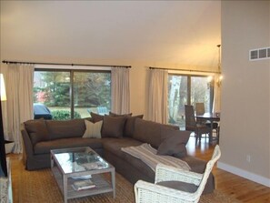 Relax in this stylish living room with a large sofa, coffee table, and wicker chair, surrounded by large windows and a dining area.