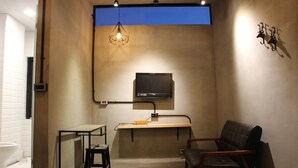 Ein kleines Zimmer mit einem an der Wand befestigten Fernseher, einem Schreibtisch mit Stuhl und einer Pendelleuchte.