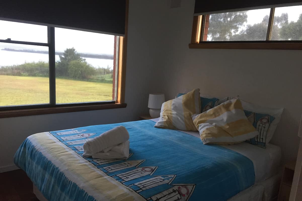Una camera da letto con un letto, cuscini, una finestra vista su un campo e una lampada su un comodino.