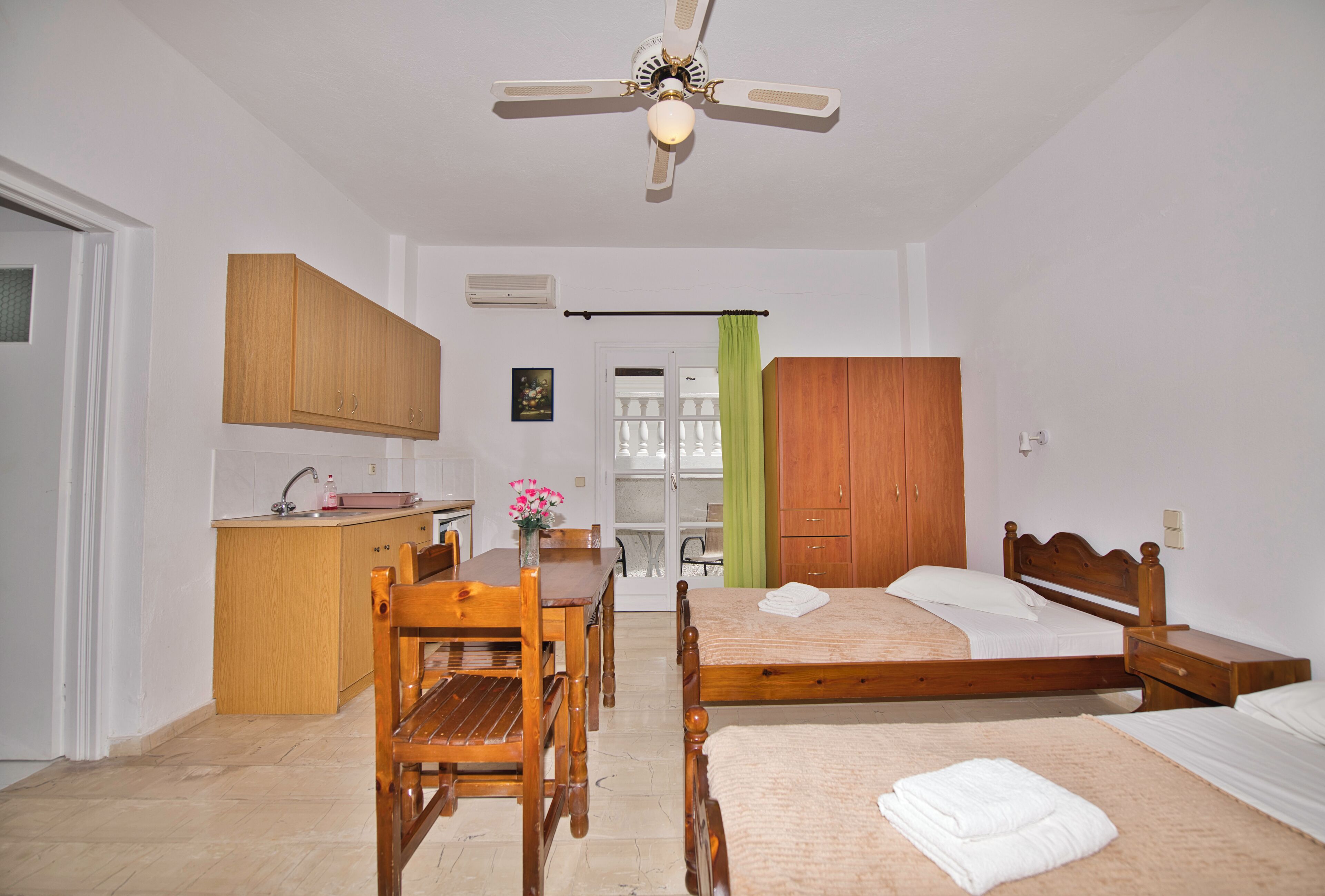 A bedroom with a bed, wooden furniture, a ceiling fan, and a small kitchenette.