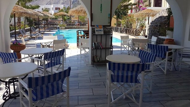 An outdoor seating area with white tables and blue-striped chairs, overlooking a swimming pool and a mountainous landscape. - Ira Apartments (Santorini)
