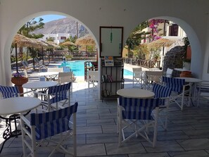 An outdoor seating area with white tables and blue-striped chairs, overlooking a swimming pool and a mountainous landscape. - Ira Apartments (Santorini)