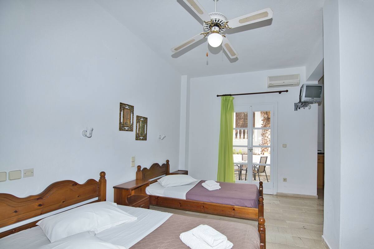 Une chambre avec deux lits en bois, un ventilateur de plafond et une vue sur un espace extérieur, accessible par une porte.