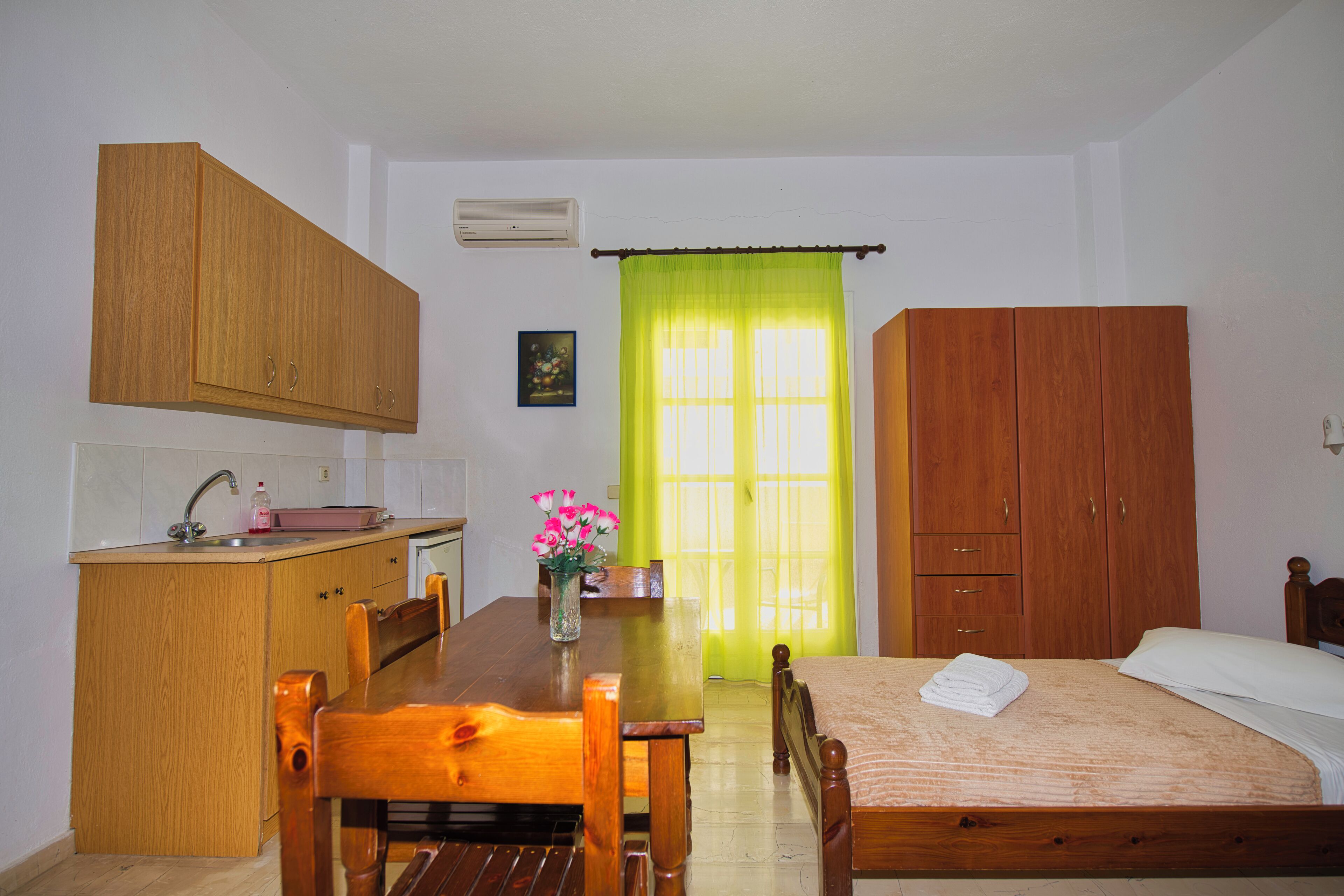 A small, clean room with a wooden bed, a wooden desk with a chair, a small kitchen area with a sink, and a window with a green curtain.