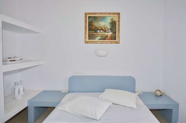 A bedroom with a bed, a nightstand, a shelf with decorative items, and a painting on the wall. - Ira Apartments (Santorini)