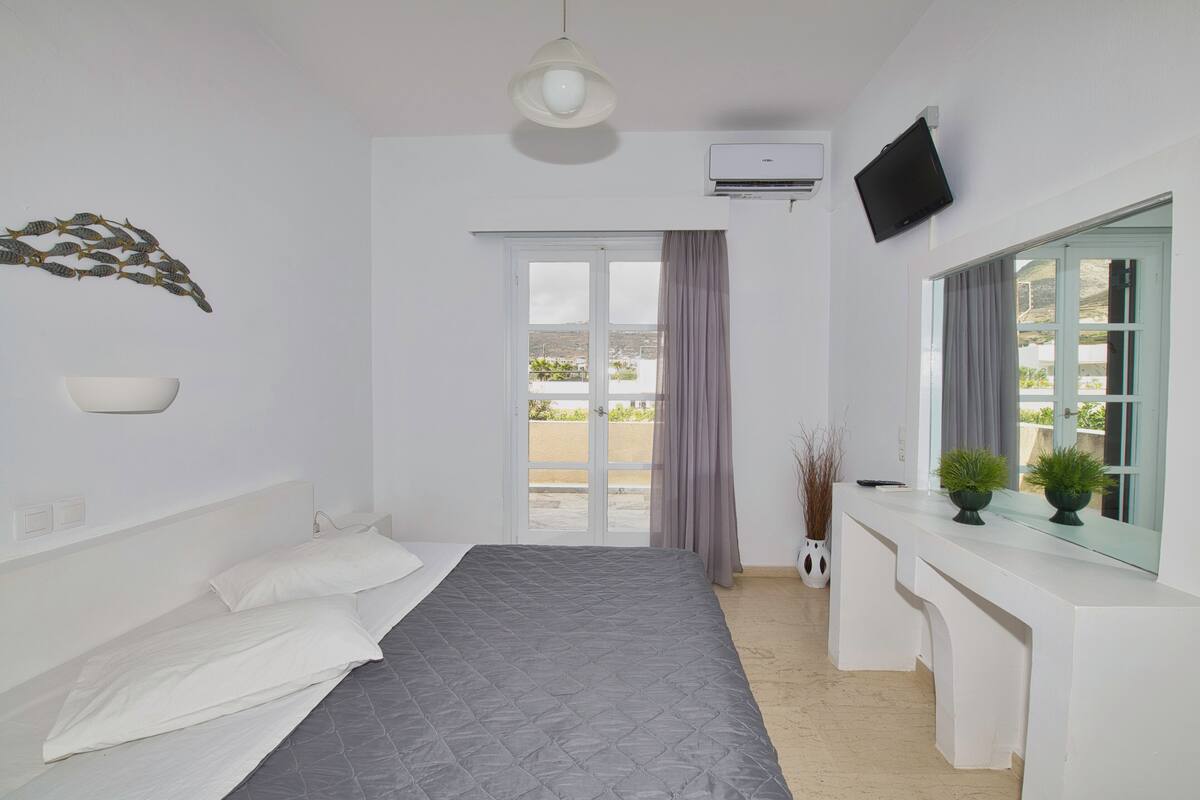 Une chambre à coucher avec un lit, un bureau, une télévision et une fenêtre avec des rideaux.