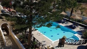 A pool area with a clear view of the sea, surrounded by trees and a white building with a stone floor. - Ira Apartments (Santorini)