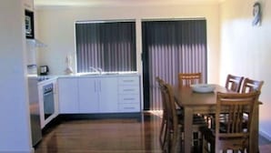 A compact kitchen with white cabinets, a dining table with chairs, and a window with curtains. - Glenaire apartments at Pontifex (Strahan)