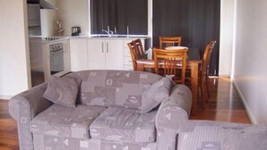 A living room with a grey patterned sofa, wooden dining table, and chairs. A kitchen with white cabinets and a stove is visible in the background. - Glenaire apartments at Pontifex (Strahan)