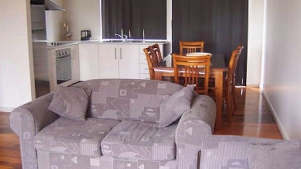 A living room with a grey patterned sofa, wooden dining table, and chairs. A kitchen with white cabinets and a stove is visible in the background.
