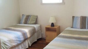 A bedroom with two single beds, a nightstand with a lamp, and a window providing natural light. - Glenaire apartments at Pontifex (Strahan)