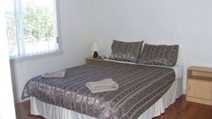 A bedroom with a bed, a nightstand, a lamp, and a window. - Glenaire apartments at Pontifex (Strahan)