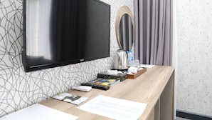 A hotel room with a flat-screen TV mounted on the wall, a wooden desk with a coffee maker, remote controls, and a magazine.