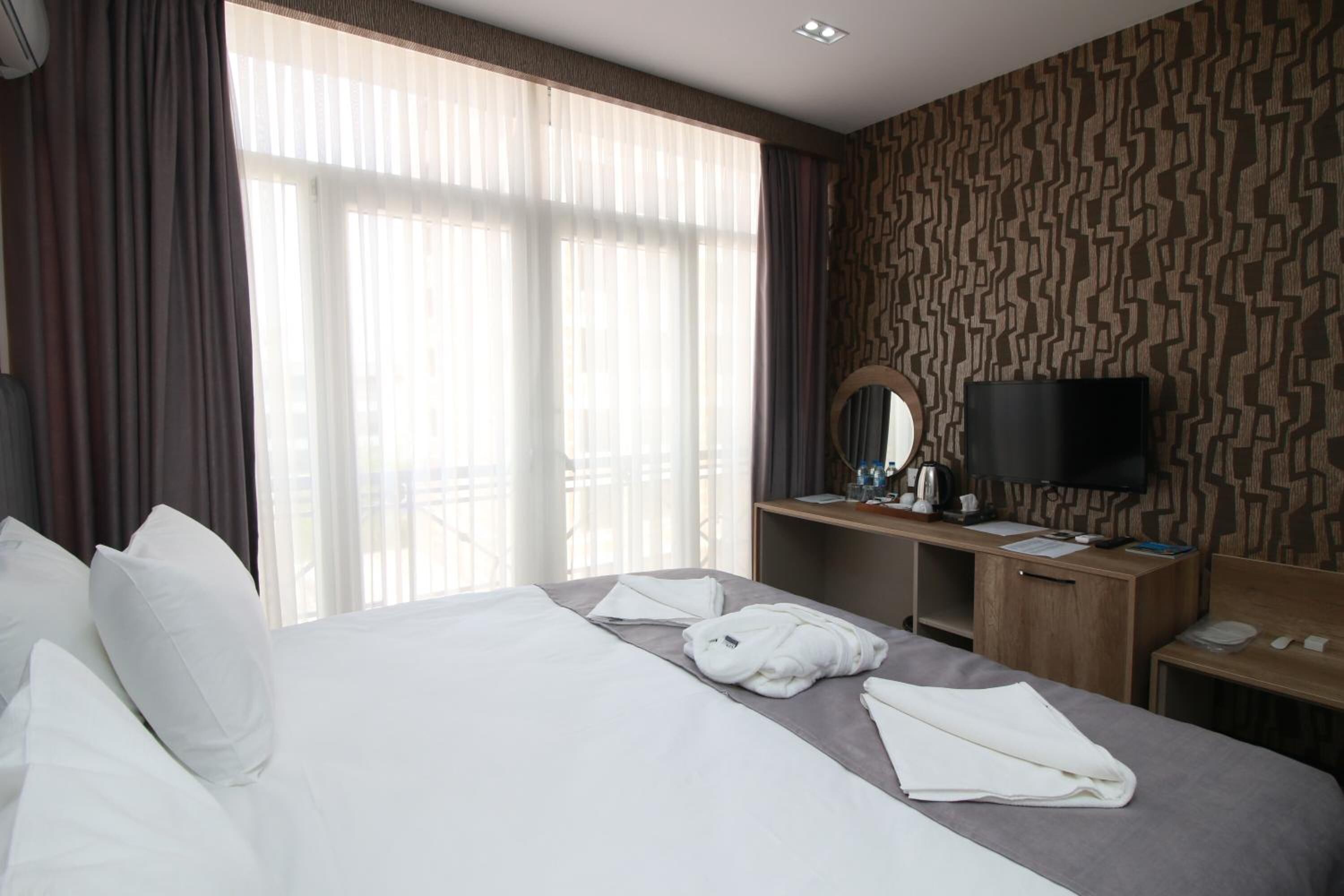 A hotel room with a large bed, a desk with a flat-screen TV, and a window with sheer curtains.