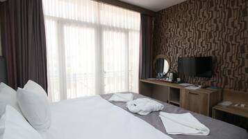 A hotel room with a large bed, a desk with a flat-screen TV, and a window with sheer curtains.