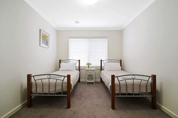A small room with two single beds, a window with blinds, a small table with a vase, and a framed picture on the wall.