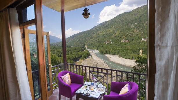A balcony with two purple chairs, a table with plates and a bottle, and a view of a river and mountains. - Regenta Inn By Riverside Manali (Kullu)