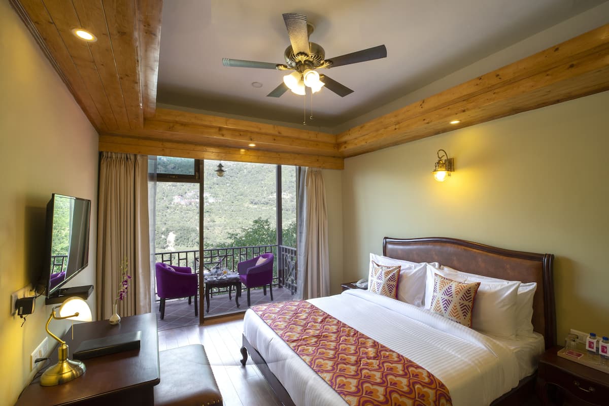 A hotel room with a large bed, a ceiling fan, a TV, a desk with a lamp, a balcony with a chair, and a view of the outdoors.