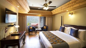 A hotel room with a large bed, a desk with a lamp, two chairs, a ceiling fan, and a TV on the wall. - Regenta Inn By Riverside Manali (Kullu)