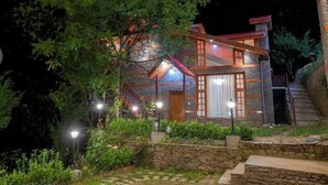A rustic cabin with a stone exterior, wooden beams, and a red roof. It features a large window with curtains and is surrounded by lush greenery. - Regenta Inn By Riverside Manali (Kullu)