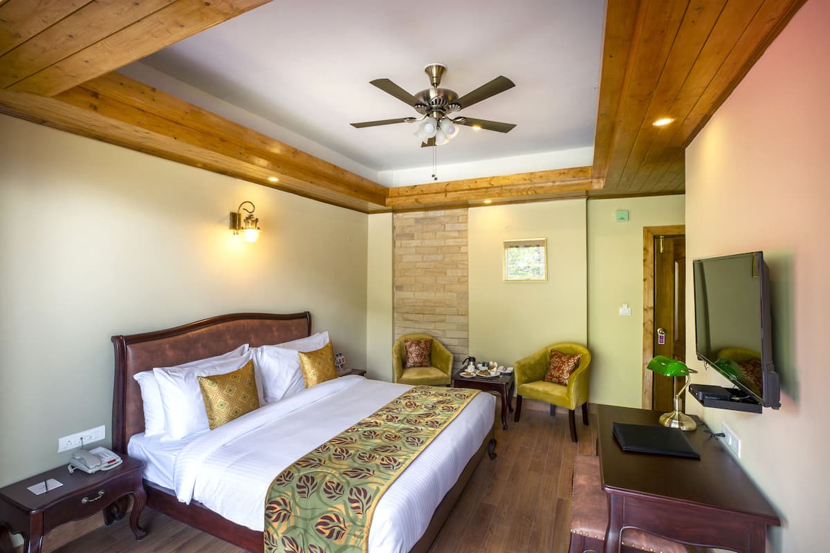 A hotel room with a wooden ceiling, a bed with a patterned bedspread, two armchairs, a desk with a laptop, and a ceiling fan.