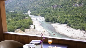A table set with a plate of food, a glass of orange juice, and a vase of flowers, with a view of a river and forested hills. - Regenta Inn By Riverside Manali (Kullu)