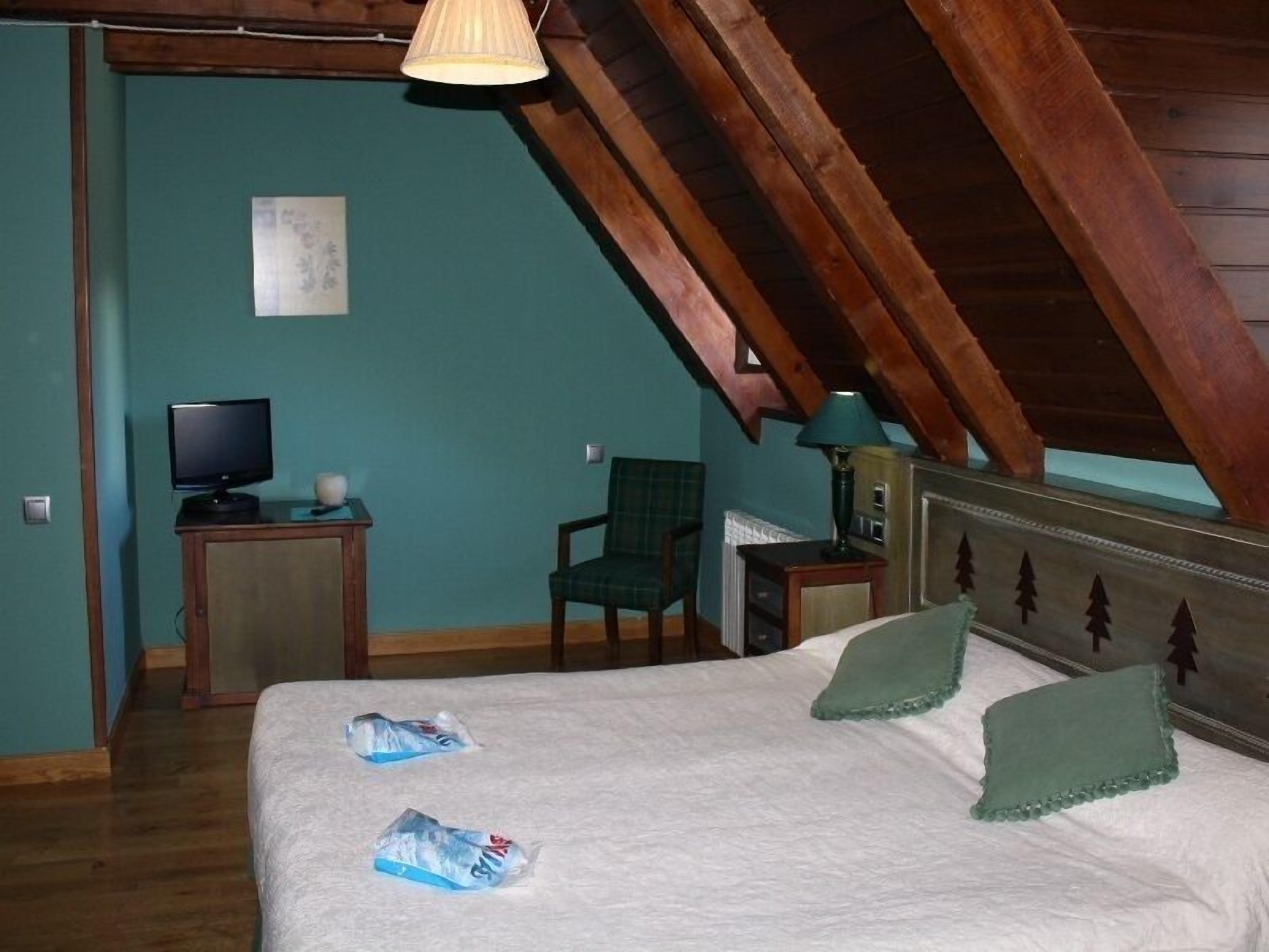 Un dormitorio con techo inclinado, una cama con almohadas verdes, un televisor sobre una mesita de noche de madera y una silla.
