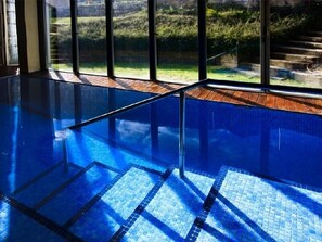 Una piscina interior con agua cristalina azul, escalones de madera y vistas a zonas verdes.