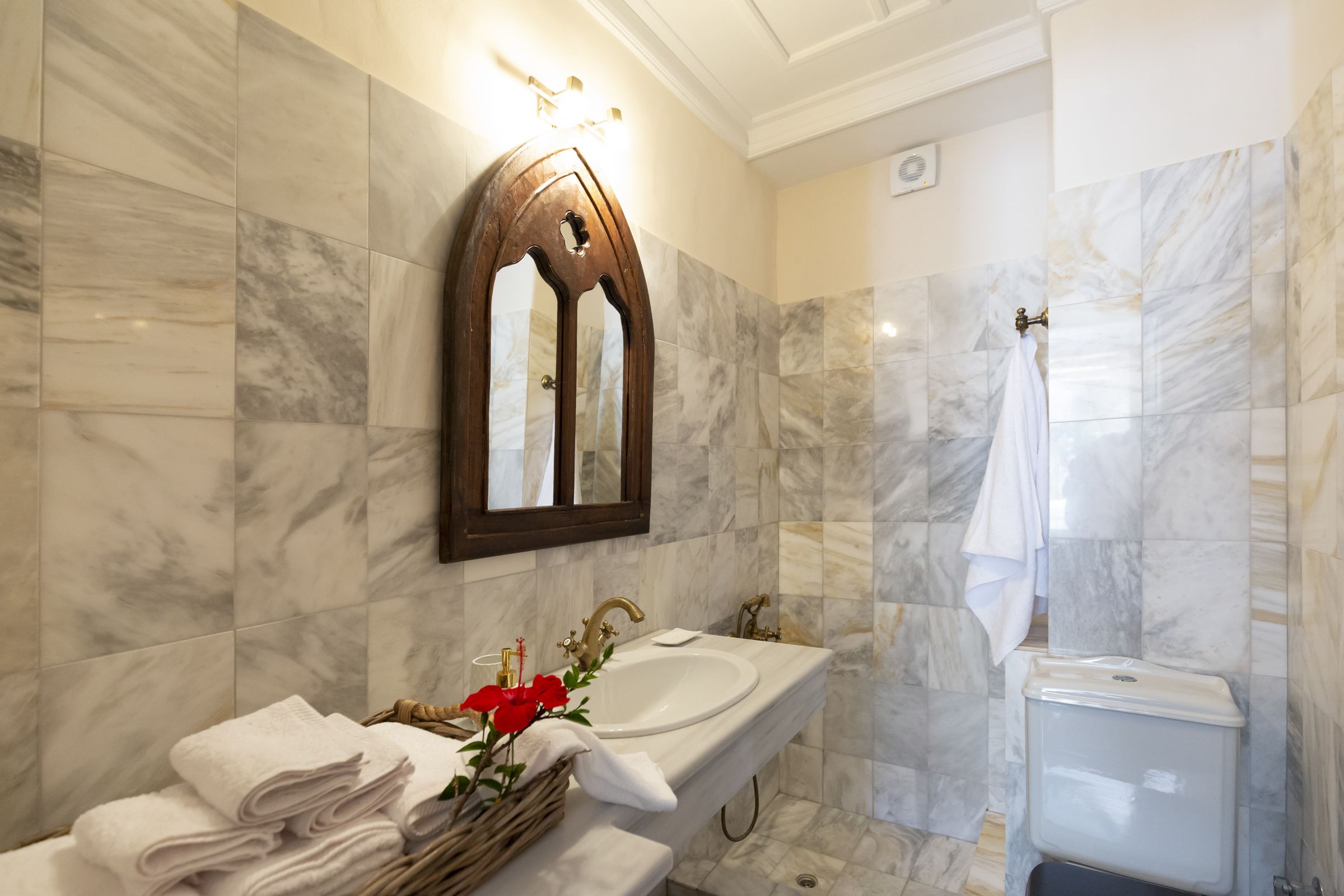 A bathroom with a marble-tiled wall, a wooden-framed mirror, a white sink, and a white toilet.