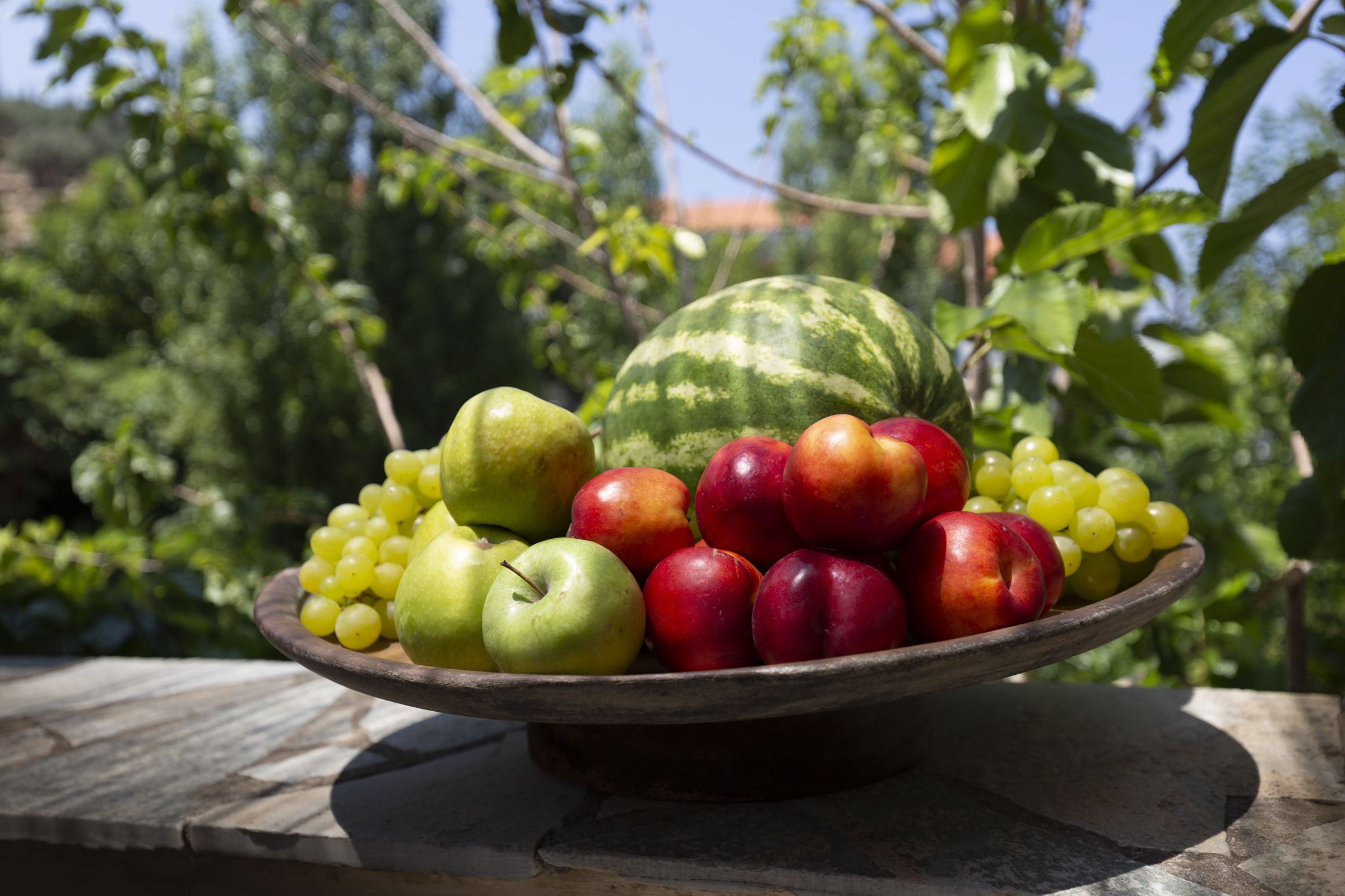A wooden bowl filled with fruits including apples, grapes, and a watermelon, placed on a stone surface with a blurred green background.