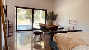 Quarto moderno com uma cama grande, área de estar com mesa e cadeiras, planta em vaso e vista para área externa por uma porta de vidro deslizante.