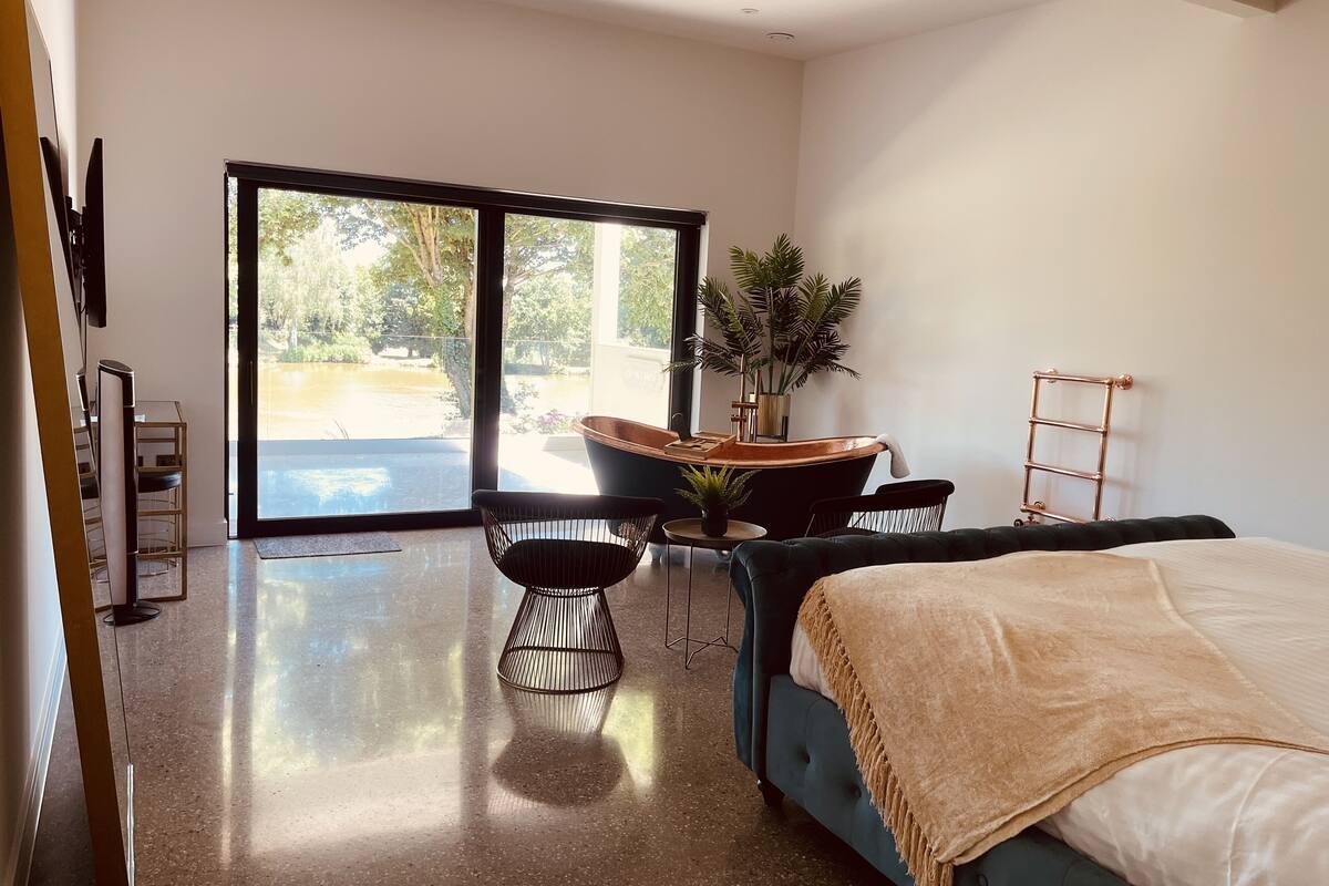A modern bedroom with a large bed, a seating area with a table and chairs, a potted plant, and a view through a sliding glass door to an outdoor area.