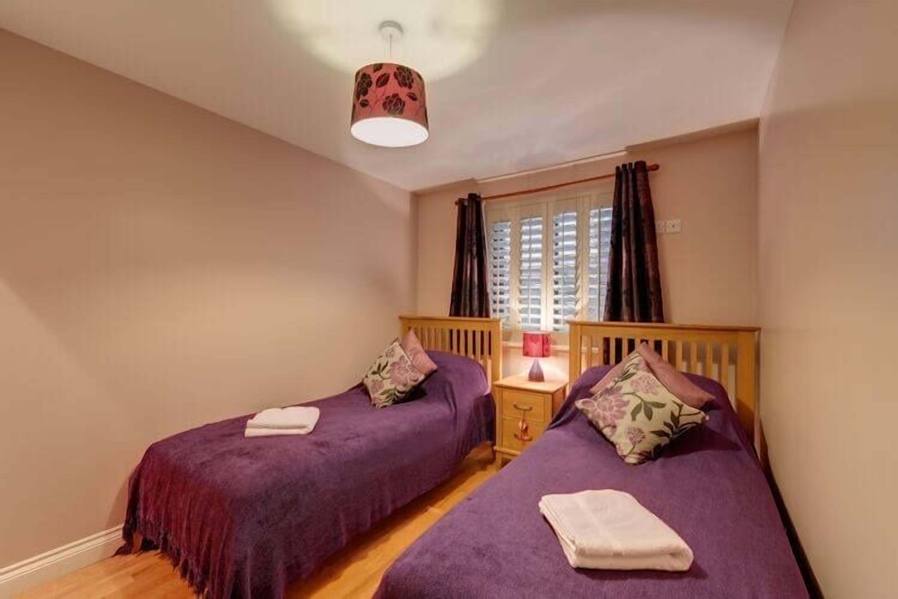 A bedroom with two single beds, wooden headboards, a bedside table with a lamp, and a window with curtains.