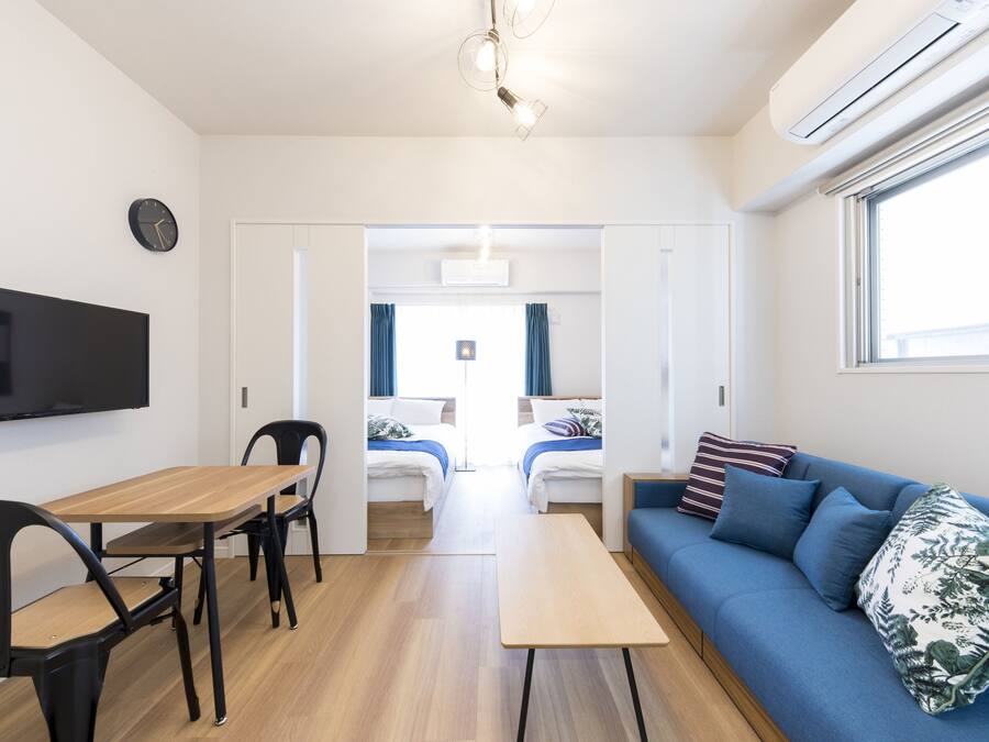 A modern room with a wooden table, two chairs, a sofa with blue cushions, a flat-screen TV, and a ceiling light fixture.