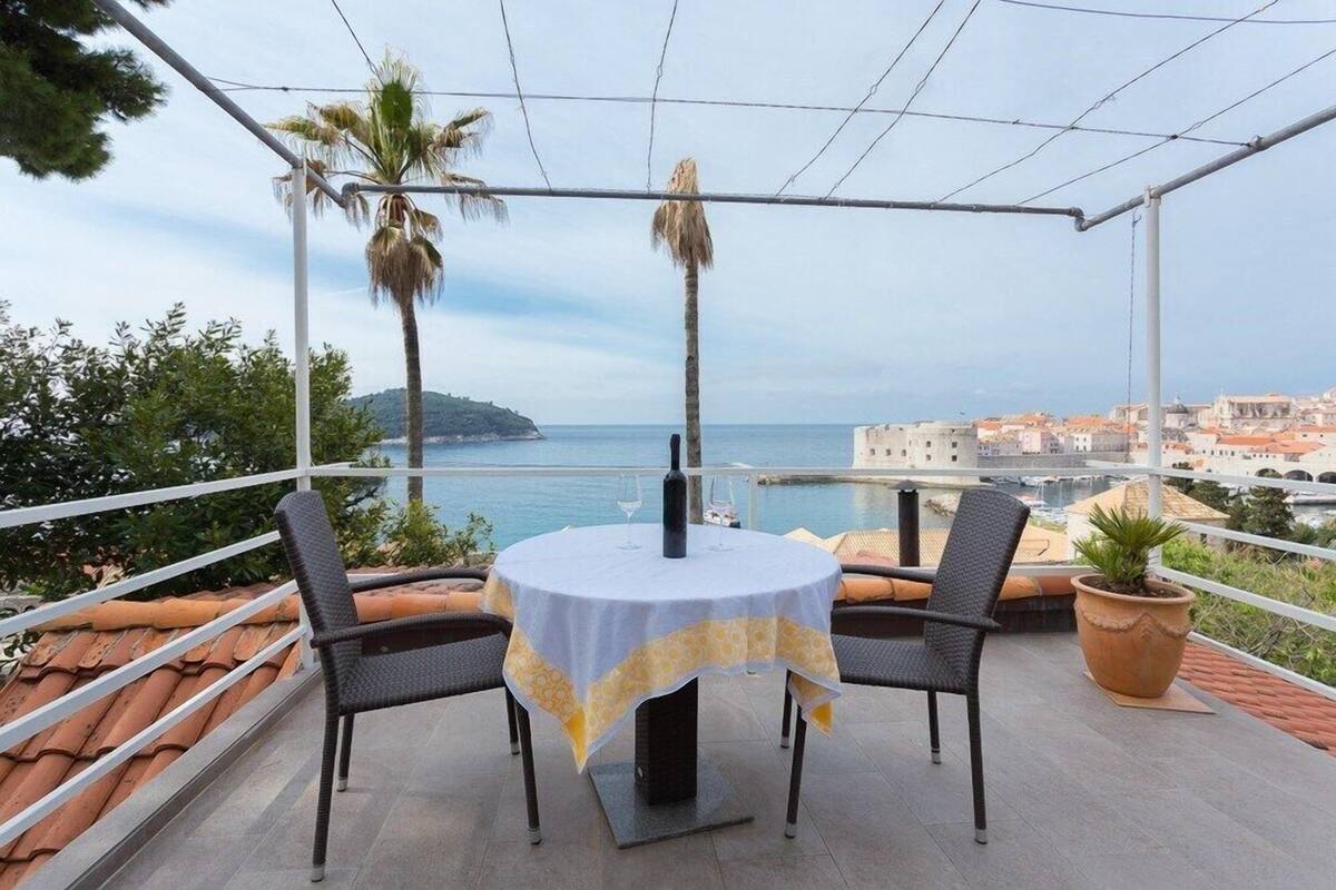 Une terrasse avec une table ronde, deux chaises, un pot de fleurs et une vue sur la mer et les bâtiments.