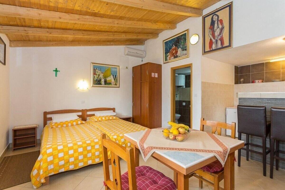 Une pièce avec deux lits, une table à manger agrémentée d’un panier de fruits, un coin cuisine et un tableau accroché au mur.