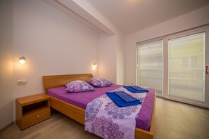 A bedroom with a wooden bed, purple bedding, a nightstand, and a window with blinds. - Apartments Emil Zora (Omis)